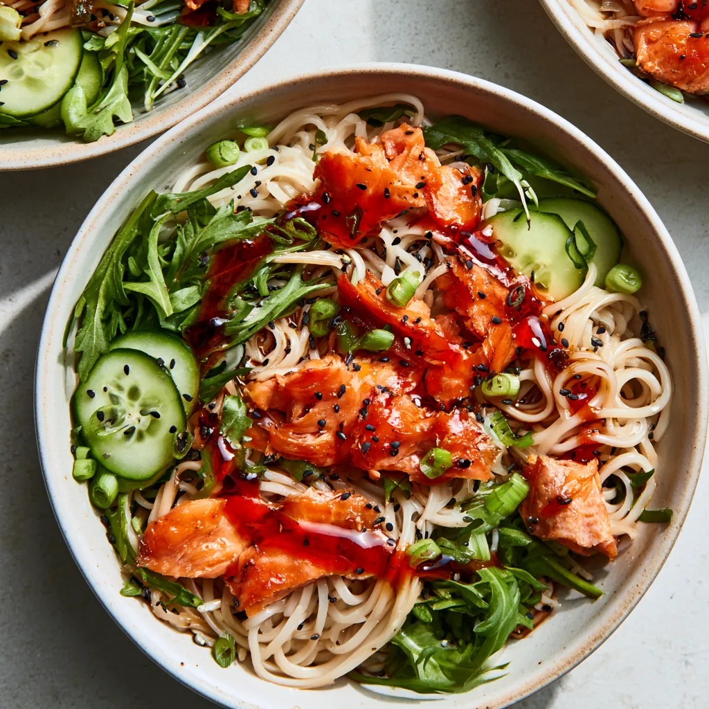 From above, two shallow bowls filled with ivory rice noodles tangled with vivid green arugula, topped with lacquered caramelized salmon flakes, half-moons of cucumber, scattered scallions, black sesame seeds, and a dramatic ruby chili-oil swirl over a gleaming soy-black-vinegar dressing.