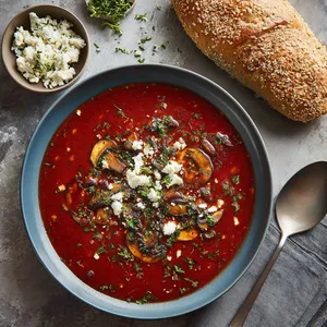The soup occupies the center of the plate showcasing bold, deep red soup dotted with chunks of Portobello mushrooms and vibrant chopped herbs. On the side, a small bowl of crumbled feta cheese and a fresh whole grain roll.