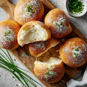 Golden-brown buns with a soft, fluffy interior, arranged on a wooden board with green chives scattered around. The buns have a dusting of flour on top, making them look rustic and homely. Half a bun is torn open to show the soft, pillowy texture inside.