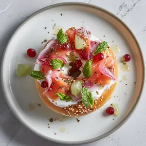 From above, a golden sesame-speckled donut half sits open-faced on a matte white plate, lavishly swirled with creamy white panna cotta ‘schmear,’ topped with silky ribbons of ruby strawberry ‘lox,’ tiny green grape ‘capers,’ translucent blush apple ‘onion,’ and delicate mint fronds. A few sesame seeds and glossy syrup dots complete the hyper-realistic New York deli look.