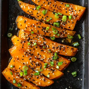 The vibrant orange hues of the BBQ glazed pumpkin wedges are beautifully contrasted by scattering of black sesame seeds and chopped fresh spring onions over each piece. Set against a rustic, charcoal-black grill tray, this dish makes for an eye-catching, appetizing image.