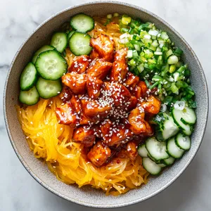 From an aerial view, behold a vibrant spectrum of colors in a beautifully presented bowl. A bed of golden spaghetti squash in the heart of the bowl forms the base, contrasted with bright, ruby-red gochujang glazed chicken pieces scattered about. There's an array of green cucumber and radish pickle slices gently placed along one side of the bowl. Chopped scallion and a sprinkling of white sesame seeds on top offer a delightful contrast.