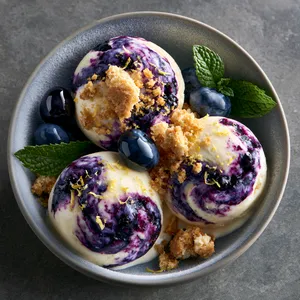 From above, a shallow frosty bowl holds three round scoops of pale ivory ice cream dramatically marbled with deep violet blueberry swirls, showered with golden cookie crumble, fine lemon zest, and a few glossy fresh blueberries and mint leaves for a vivid, elegant contrast.