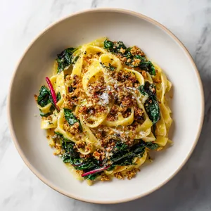 From above, a shallow bowl of glossy tagliatelle twirled into a loose nest, streaked with emerald chard ribbons and ruby chard stems, finished with golden walnut crumble, tiny lemon zest threads, black pepper, and a light shower of grated cheese over a warm ivory plate.