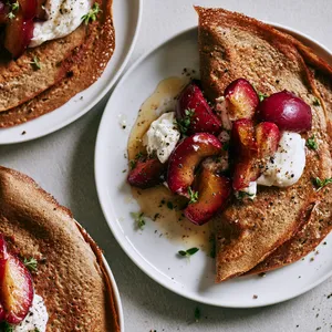 From above, two deep golden buckwheat crêpes sit loosely folded on white plates, their edges lacy and rustic. Glossy ruby-purple roasted plum wedges spill from the center beside clouds of whipped goat cheese, scattered thyme leaves, cracked black pepper, and a light honey sheen, with a few tiny greens for contrast.