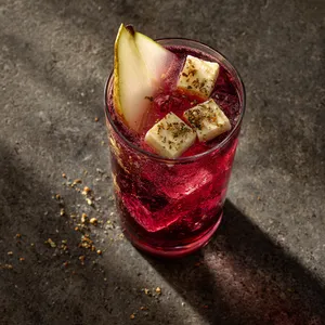 An inviting cocktail in a clear highball glass with deep ruby color from infused Swiss Chard. The pear slice resting on the rim gives a subtle contrast. Floating Ardennes cheese cubes and a sprinkle of nutmeg on the surface adds a gourmet finish.