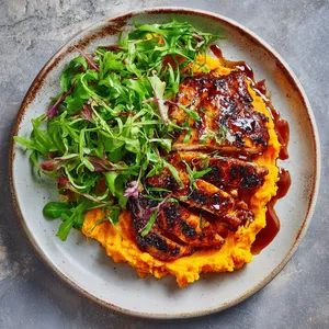 The glossy BBQ chicken lies atop a bed of vibrant green watercress, which contrasts sharply with the sunset orange of the sweet potato mash. The whole plate is packed with vivid colors and dynamic textures, looking as tempting as it tastes.