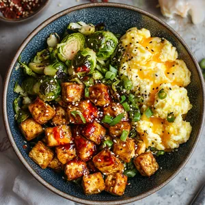 A vibrant bowl, overflowing with warm charred brussels sprouts, ruby-red Asian chili sauce drizzle, and succulent amber-hued cubes of smoked tofu. Flecks of sesame seeds and shredded green onions add bursts of color. A few fluffy scrambled eggs rest at the side.