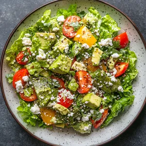 A riot of colors covers the plate, delicately charred pieces of green nopales scattered amongst vibrant chunks of red bell pepper and tomatoes, sprinkled with flecks of white crumble cheese, all resting on a bed of crisp romaine lettuce. Drizzled over all this is a creamy, avocado-based dressing that lends an appealing sheen to the ingredients.