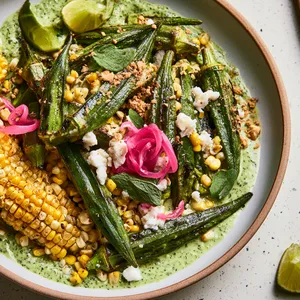 From above, a wide shallow plate filled with glossy charred okra and golden corn, dotted with ivory cheese, ruby pickled onion curls, and emerald oregano leaves over a pale green herb sauce, finished with toasted cassava crumbs and lime wedges.