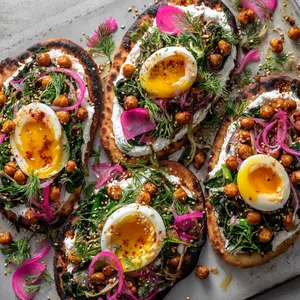 From above, four oval flatbreads are spread with snowy white labneh and topped with deep green charred ramps, halved jammy eggs with glowing golden centers, crispy paprika-sumac chickpeas, bright herbs, pink pickled onions, and a glossy drizzle of olive oil and honey, finished with scattered sesame and crimson Aleppo pepper.