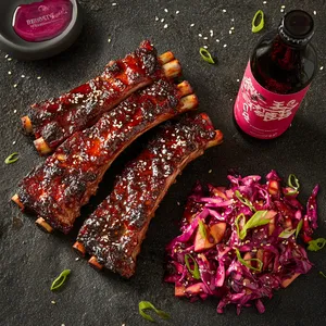 Hearty racks of ribs glistening under a glossy, deep reddish-brown glaze, charred tips peeking out. A generous heap of bright magenta slaw sits by, dotted with vibrant green spring onions and sesame seeds. Bottles of icy drink wink in the late afternoon sun.