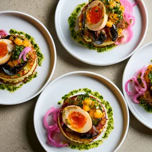 From above, four plates show golden lacy cassava cakes topped with glossy folds of roasted eggplant, halved jammy eggs with deep orange centers, jewel-like mango relish, emerald herbs, pink pickled onion curls, and a drizzle of green herb oil on bright white plates.