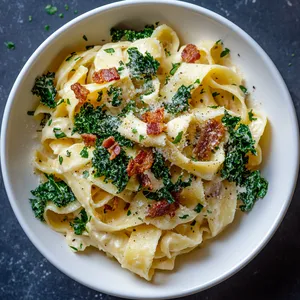 From above, evenly coated cream-coloured pasta ribbons are seen, speckled with vibrant green pieces of crispy kale and crispy reddish-brown bacon crumble. The pasta is placed into a deep white pasta bowl with an aesthetic scattering of fresh parsley and a drizzle of extra olive oil on top.