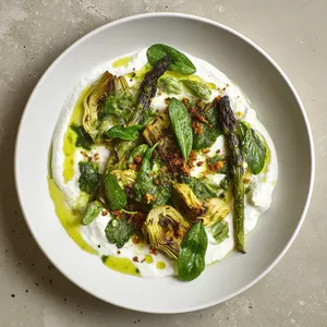 From above, a wide white plate holds a swoosh of pale cannellini cream topped with charred asparagus spears and golden roasted baby artichokes, torn burrata nestled between them, glossy sorrel ribbons and basil leaves scattered throughout, all finished with lemon zest, emerald olive oil droplets, toasted pangrattato, and a few cracked black pepper flecks for contrast.