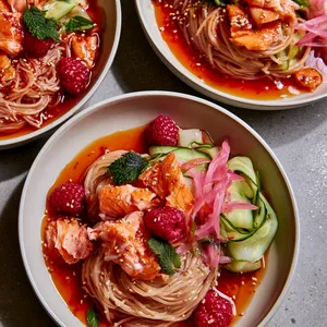 From above, wide shallow bowls hold glossy chilled noodles swirled into neat nests, topped with lacquered golden salmon flakes, ruby raspberries, pale green cucumber ribbons, pink pickled onion, scattered sesame seeds, and small leaves of perilla and mint over a vivid red-orange dressing.