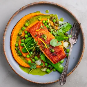 From above, a swoosh of vivid orange carrot purée anchors the plate, topped with a golden crisp-skinned salmon fillet. Bright green peas and watercress are scattered artfully around, with glossy capers, feathery tarragon leaves, and tiny shallot slices catching the light. A spooned ring of emerald-flecked vinaigrette and a few lemon zest strands make the dish look fresh, modern, and striking.
