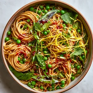 From above, a shallow bowl of glossy wheat noodles is twirled into loose nests, dotted with bright green peas and charred snap pea halves, streaked with red chili oil, scattered with sesame seeds and sliced scallions, and finished with ribbons of lemon zest and fresh mint for vivid contrast.