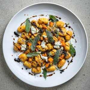 Uniform, golden-brown gnocchi pillows are lightly scattered across the plate mixed among vibrant green sage leaves and blushing butternut squash pieces. Dots of melted Gorgonzola artfully splattered around lend an unexpected pop of white while a dusting of parmesan and a drizzle of Balsamic reduction adorning the top add the final touch.
