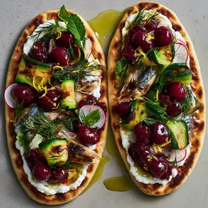 From above, two oval golden flatbreads are swirled with white whipped feta, topped with bronzed mackerel flakes, ribboned charred zucchini, glossy deep-red cherry relish, scattered dill and mint, thin radish slices, and lemon zest, finished with a shimmer of olive oil.