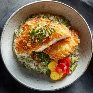 A beautiful, deep golden-brown piece of halibut tempura sits invitingly atop a cozy mound of light green spring onion rice. Colorful Japanese pickles - shades of red and yellow - lay on the side, contrasted against the pristine white ceramic plate. The combination is visually striking against the backdrop of a dark linen tablecloth.