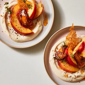 From above, two shallow dessert plates each hold a swoosh of pale crème fraîche topped with blushing peach wedges in a loose fan, jewel-like amber shiitake slices glistening between them, and delicate golden sesame tuile shards standing upright for height. Tiny thyme leaves, a thread of honey syrup, and a few toasted sesame seeds create a refined, airy, editorial look.