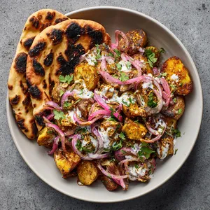 Golden, crispy sunchokes sit atop a vibrant mix of red onions, green cilantro, and white yogurt, with dark mustard seeds speckled throughout. A couple of well-grilled Tandoori masala naan rest alongside this vivacious medley, their charred edges creating a beautiful contrast.