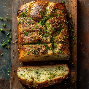 The baked loaf boasts a beautiful, burnished golden-brown crust, dusted with a sprinkling of cumin seeds interspersed with tiny specks of vibrant green chives. A neat horizontal cut reveals the fluffy, light interior of the bread, inviting and delicious.