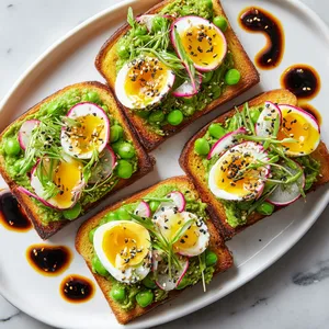 From above, four thick slices of golden toast are spread with bright green pea-edamame smash, topped with halved jammy eggs, pale pink radish ribbons, black sesame seeds, scallions, and tiny herb leaves, arranged on a white platter with glossy chili oil swirls.