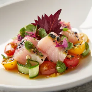 A vibrant palette of color awaits you atop a pristine white plate. Slices of yellowtail possess a slight translucence and a soft pink hue, adorned with deep green ribbons of cucumber and bright red and orange patches of heirloom tomatoes. Ground wasabi peas add bursts of jade amidst dainty flakes of sea salt. The dish is garnished with deep red shiso leaves and a sprinkle of black sesame seeds, creating an artful display that excites the eyes.