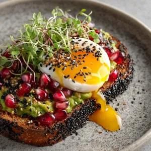 Imagine a dish emanating rustic elegance with juicy pomegranate seeds glistening like rare gems atop a luscious landscape of light green avocado, while a runny yolk from the Onsen Tamago seductively cascades over the dish. It's finished with a bright pop of microgreens and striking black sesame seeds, laying on a crusty sourdough slice.