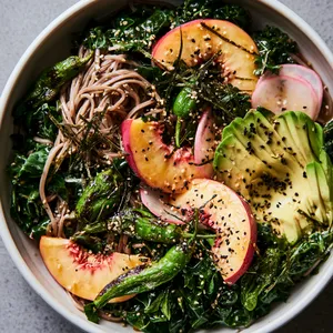 From above, a wide shallow bowl filled with glossy buckwheat soba and ribboned deep-green kale, dotted with charred green shishito peppers, crescent slices of blush-gold nectarines, fanned avocado, pale pink radish slices, black and white sesame seeds, and delicate strips of nori, all lightly gleaming with dressing.