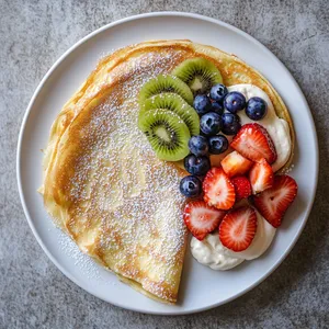 From above you can see a golden circular crêpe folded into a triangle on a white plate. The cream cheese filling peaks invitingly at the edges, and a rainbow of fruits; strawberries, blueberries, kiwi slices artistically displayed on top alongside a generous sprinkle of powdered sugar that gives a magical frosty look.