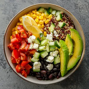 A colorful medley of diced tomatoes, cucumber, and corn against a contrasting base of black beans and white quinoa, with a final sprinkling of crumbled feta cheese on top. Lemon wedges and avocado slices artfully placed on the side further enrich the palette with their bright yellows and greens.