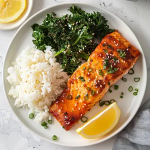 Golden glazed salmon fillet on a simple white plate, sprinkled with toasted sesame seeds. A well-seasoned pile of crispy, slightly charred kale sits next to the salmon adding a vibrant green pop to the plate. A side of short-grain white rice and lemon wedges complete the picture.
