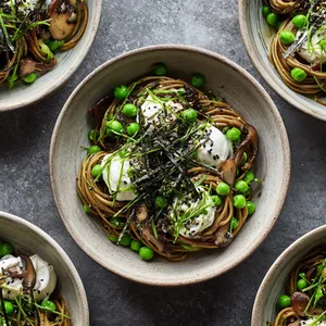 From above, a shallow bowl of taupe soba noodles twirled into glossy nests, dotted with bright green peas and browned mushrooms, with soft swooshes of whipped ricotta, a shower of finely cut chives, black sesame, thin nori ribbons, and lemon zest for a clean, modern contrast.