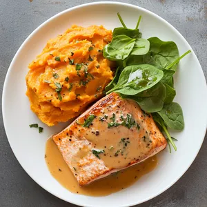 A beautiful blend of colors on a white plate: succulent pink salmon topped with a creamy, pale sauce next to a mound of vibrant orange sweet potato mash. A sprinkle of fresh green spinach leaves for garnish provides a refreshing contrast.