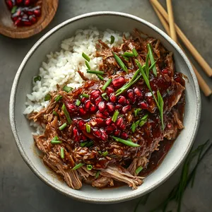 A deep, round white ceramic bowl is filled with a generous helping of slow-cooked pulled pork, glistening with a sweet and spicy pomegranate gochujang sauce. The meat is topped with bright crimson pomegranate arils and fresh green chives, alongside a side of fluffy white rice. Next to it, a pair of clean silver chopsticks rest on an earthy wooden chopstick rest.