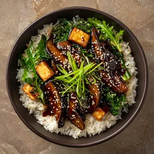 A glossy, dark teriyaki sauce coats thick slices of grilled portobello, golden tofu cubes, and vibrant greens, all nestled on a bed of fluffy white rice in a deep bowl. Upon garnishing with spring onions and sesame seeds, the dish transforms into a perfect autumn palette with bursts of green and ivory.