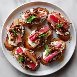 From above, six toasted crostini are arranged in a loose circle on a matte white plate, each topped with a swoosh of whipped goat cheese, glossy blush-pink rhubarb batons, chopped candied pecans, and tiny mint leaves, finished with a glistening amber drizzle and cracked black pepper.