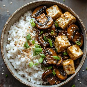 The vibrant hues of caramelized onions, garlic, and mushrooms glisten in a glossy sauce sprinkled with sesame seeds. Plump cubes of tofu add contrasting shades of white. The dish is garnished with green spring onion rings scattered casually on top. All served on a backdrop of fluffy brown Jasmine rice in a rustic bowl.