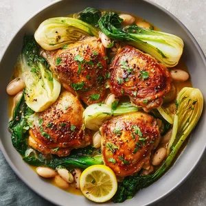 From above, golden bronzed chicken thighs rest over creamy white beans, pale green bok choy, and soft fennel in a glossy amber sauce, finished with chopped parsley, cracked pepper, and thin lemon slices for a bright, photogenic contrast.