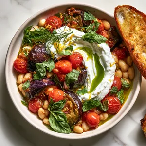 From above, a shallow bowl of glossy aubergine-purple eggplant and ruby cherry tomatoes nestled among creamy white beans, topped with a swoosh of white whipped ricotta, torn basil and mint, fennel fronds, lemon zest, and a drizzle of emerald olive oil, with toasted bread angled alongside.