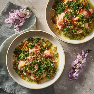 Two deep bowls filled with smoked-pink Sakura chicken under a garnish of vibrant green finely chopped parsley. The chicken and vegetables sit on top of a creamy, light broth. The dish is accented with fine blossom petals, which lend the dish its appeal and sophistication.