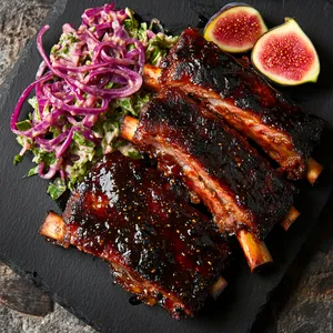 On a black stone plate, two glossy, deep-red ribs rest on a bed of vibrant green cilantro lime coleslaw, glistening with the sticky fig-infused BBQ sauce. A side of grilled figs cut in halves, with dark grill marks on them, sit on the edge of the plate, the vibrant inner flesh contrasts with the charred outer layer. A sprinkling of vibrant pink pickled red onions is scattered across the entire dish, providing a pop of color.