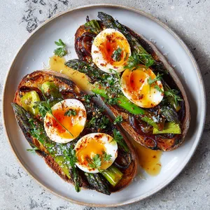 From above, two rustic toasts sit on a light plate, piled with glossy paprika-tinted onions and charred green asparagus tips, each topped with halved jammy eggs. A swipe of tomato beneath peeks out at the edges, finished with tiny parsley leaves, flaky salt, black pepper, and a shimmering drizzle of olive oil.