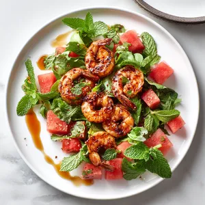 A vibrant array of Sriracha-glazed pink shrimp, juicy watermelon cubes and bright greens artfully arranged on a white dinner plate. Garnished with fresh mint leaves and a drizzle of honey glaze, making this dish a feast for the eyes.