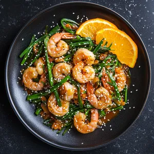 At the center of a sleek black plate, a pile of glossy orange-colored stir-fry catches the eyes, speckled with green chive blossoms and pink shrimp. Slices of orange adorn the side of the plate, suggesting a sweet kick, while a sprinkle of white sesame seeds over the colorful mound adds a final touch of contrast.