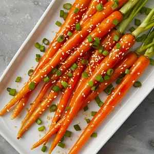 Slender, glossy carrot spears in varying sunset hues of orange, laid elegantly across a minimalistic white platter. The dish is generously sprinkled with toasted white sesame seeds and flecks of chopped spring onion greens.