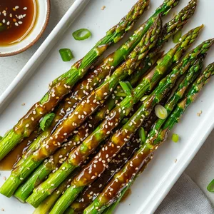 Golden-brown Asparagus skewers lay perfect on a clean-white oblong porcelain platter, delicate sesame seeds scattered atop, providing a delightful contrast. A small bowl holding extra glaze sits on the corner, shimmering in the soft light. The vibrancy of green asparagus, the caramel tones of the glaze, and the rustic texture of sesame seeds make this dish a vibrant, color-filled visual feast.